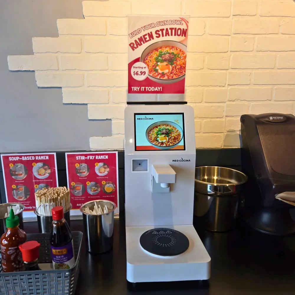 NEO CUCINA ramen cooker on a countertop in a boba shop, surrounded by cups, ingredients, and condiments, with digital display showing ramen preparation.