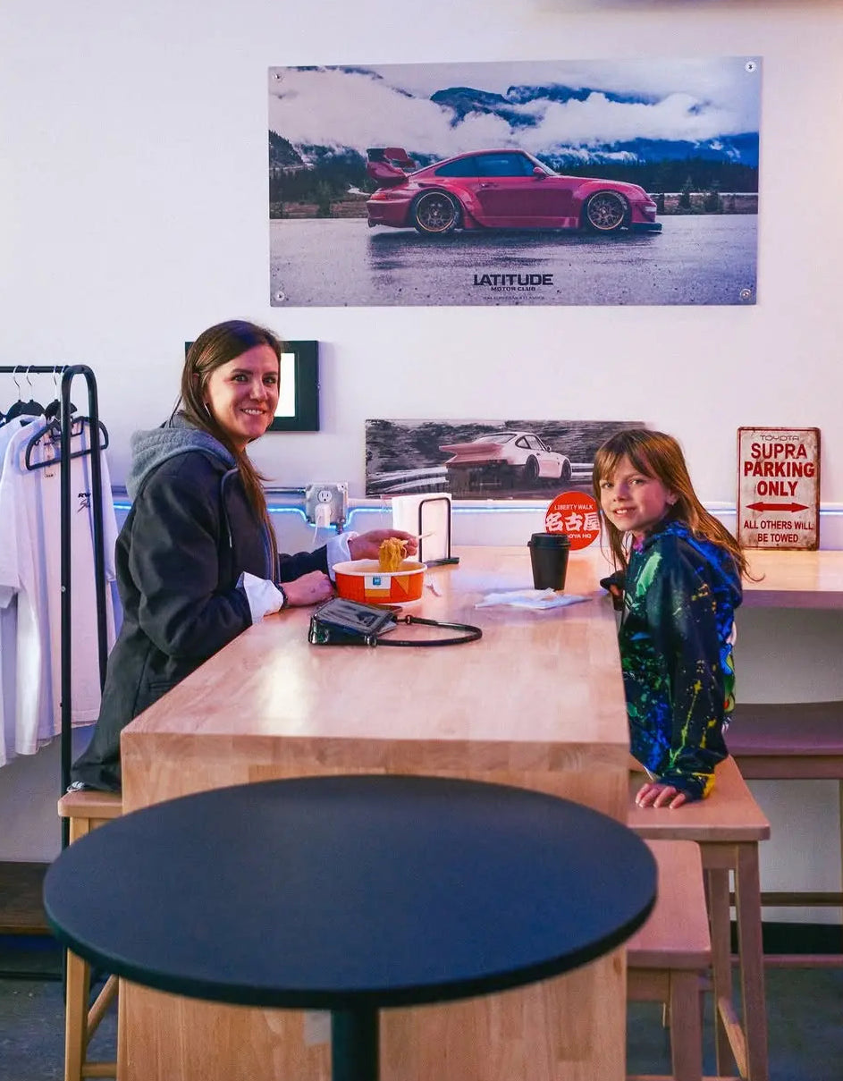 A mother and child enjoying hot ramen prepared by a NEO CUCINA smart cooker in a car-themed cafe or garage lounge. The image showcases the family-friendly appeal and versatility of automated self-serve ramen stations in non-traditional food service settings.
