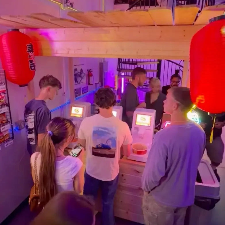 A crowd of young customers using NEO CUCINA smart commercial ramen machines in a vibrant, neon-lit entertainment venue. The image demonstrates the high engagement and ease of use of the self-serve cooking stations in a busy, themed restaurant environment.