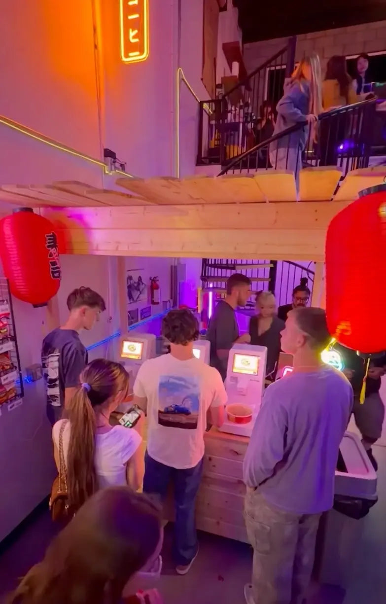 A crowd of young customers using NEO CUCINA smart commercial ramen machines in a vibrant, neon-lit entertainment venue. The image demonstrates the high engagement and ease of use of the self-serve cooking stations in a busy, themed restaurant environment.