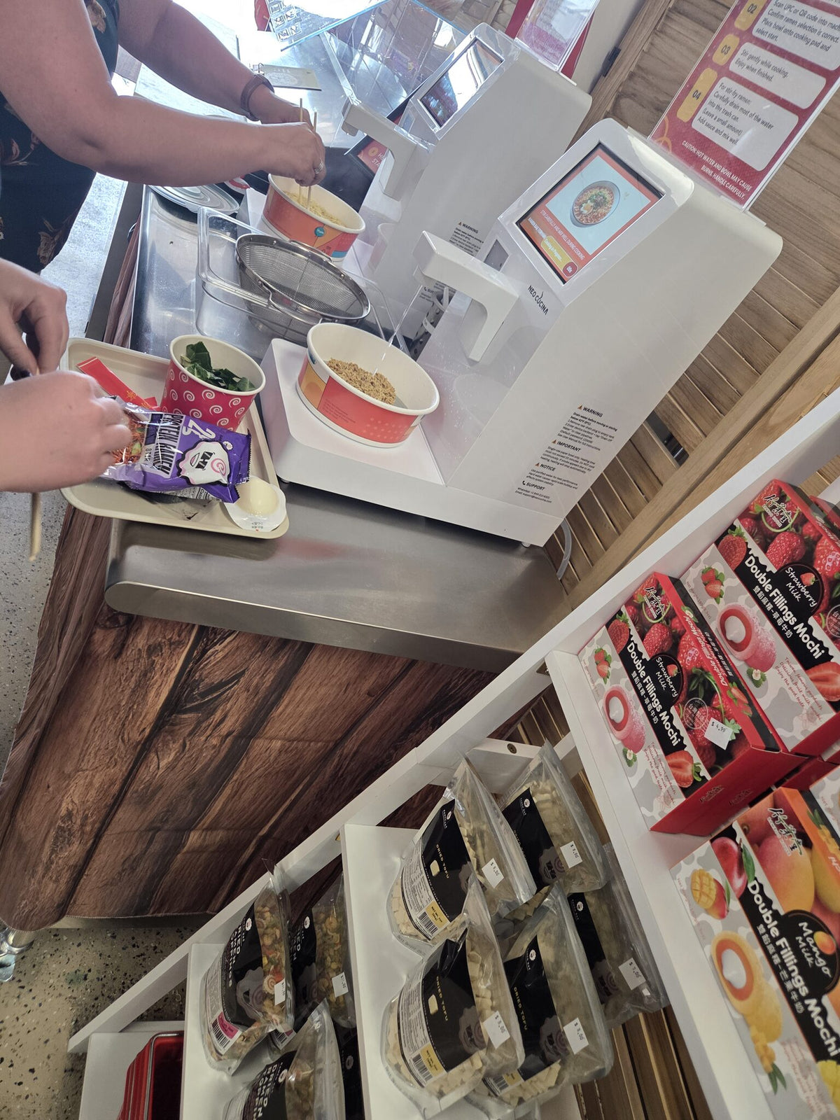 Customers cooking ramen at two machines with topping trays, Helena meal-prep shop